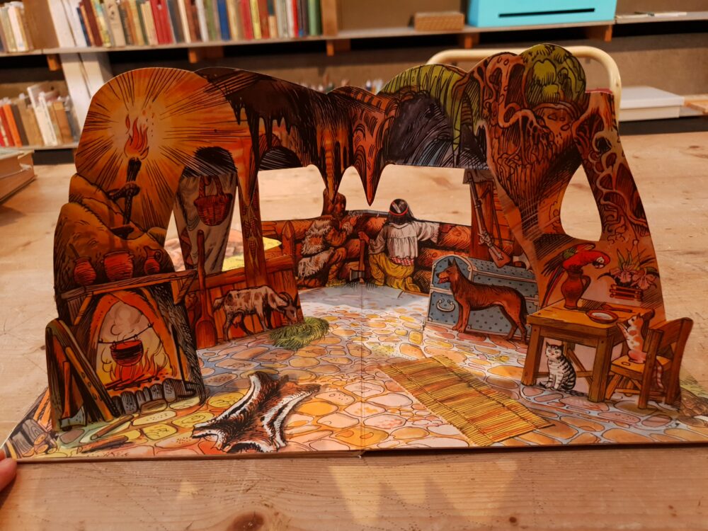 The 3D pop-up book features a double page of Crusoe’s home, showing a table, fireplace, cave, and multiple animals.